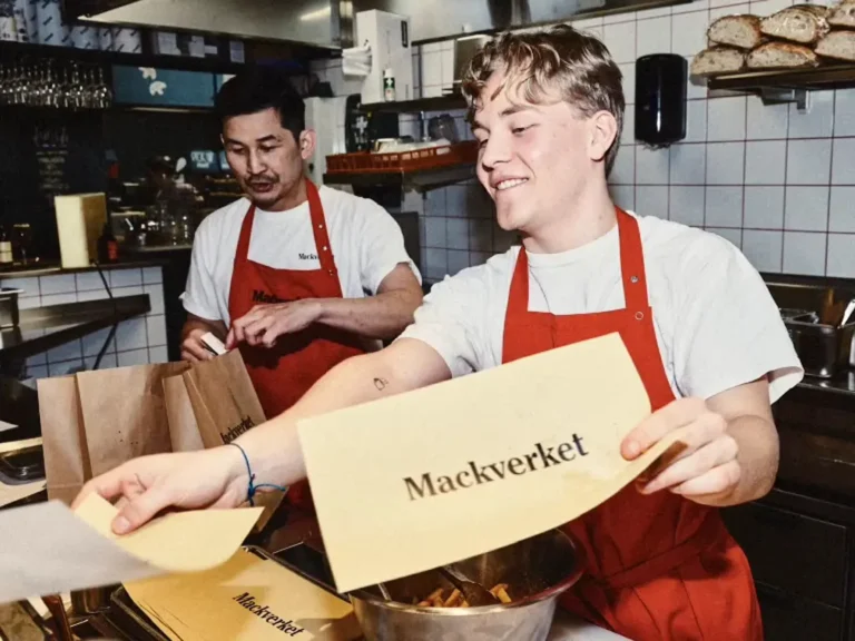 Två kockar i röda förkläden arbetar i Mackverkets kök. En yngre man i förgrunden ler och räcker över papper med Mackverket-logotypen, medan en kollega packar papperskassar i bakgrunden.