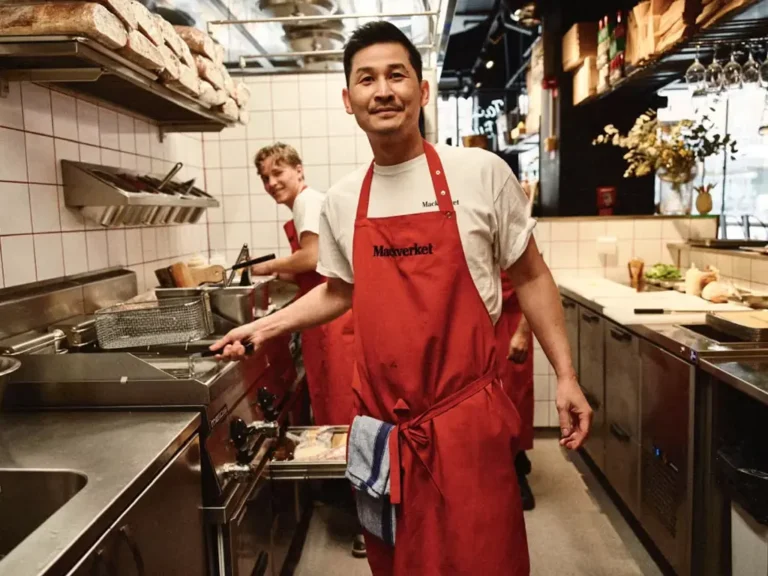 Kock i rött Mackverket-förkläde ler i köket, med kollegor i bakgrunden.