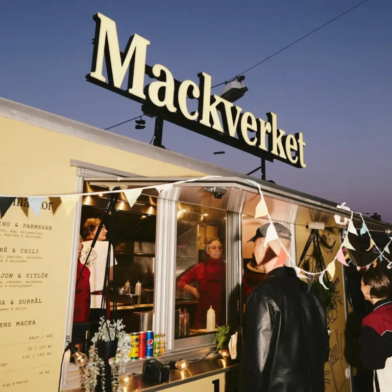 Mackverkets food truck i skymningen med en stor belyst logotyp på taket och kunder vid serveringsluckan.