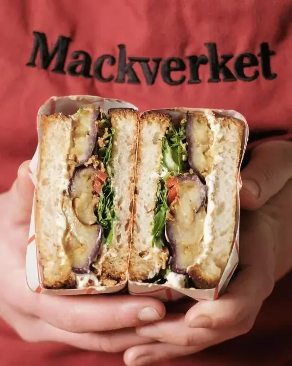 Två händer håller upp en halverad varm macka framför ett rött Mackverket-förkläde.