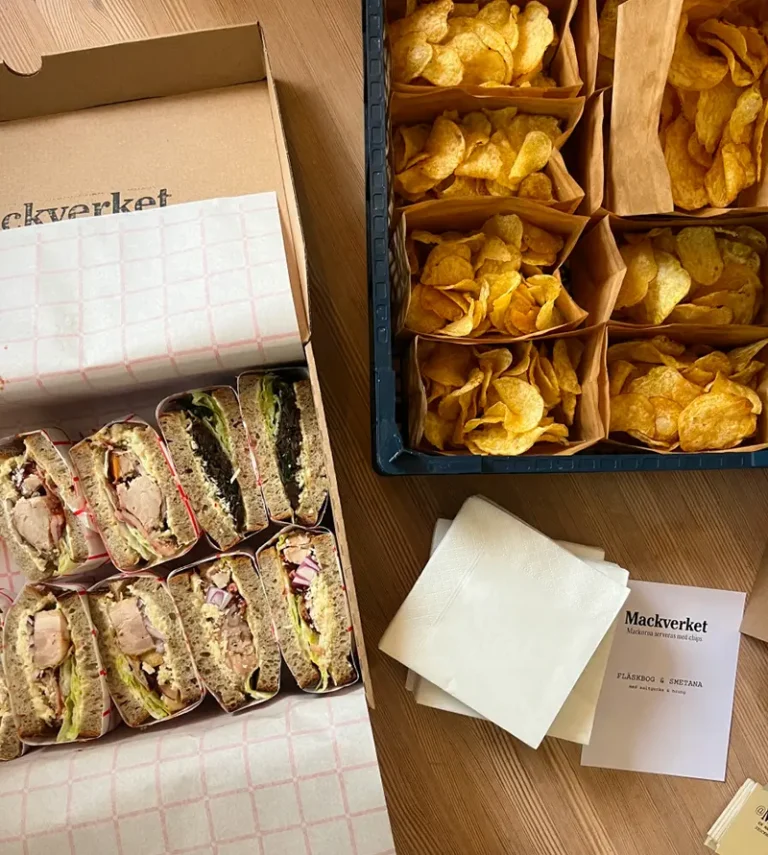 En cateringbox fylld med halverade mackor bredvid påsar med chips och servetter från Mackverket.