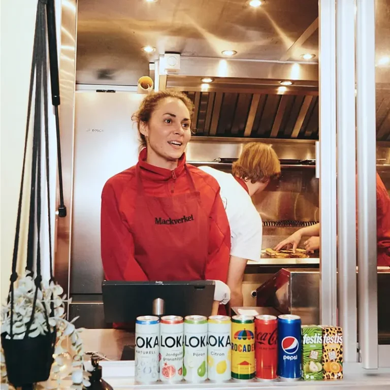 Kvinna i rött Mackverket-förkläde står vid kassan i en food truck med drycker uppradade i förgrunden.